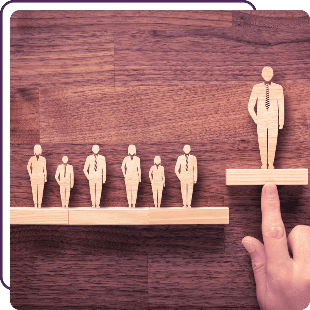 Wooden figures representing people stand on blocks of increasing height on a wooden surface. A hand elevates the tallest block with one figure, suggesting leadership or success.